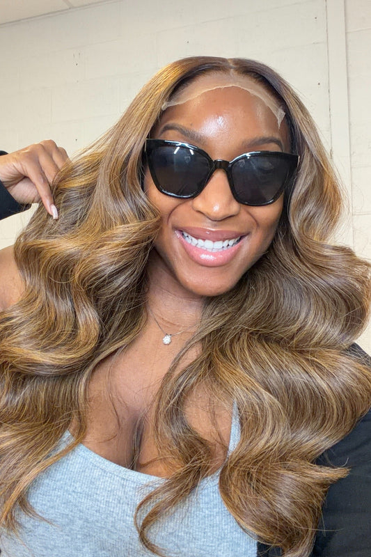 A smiling woman wearing a long, wavy, highlighted wig with a clear lace front, black sunglasses, and a gray top, with a hand adjusting the hair on her shoulder.